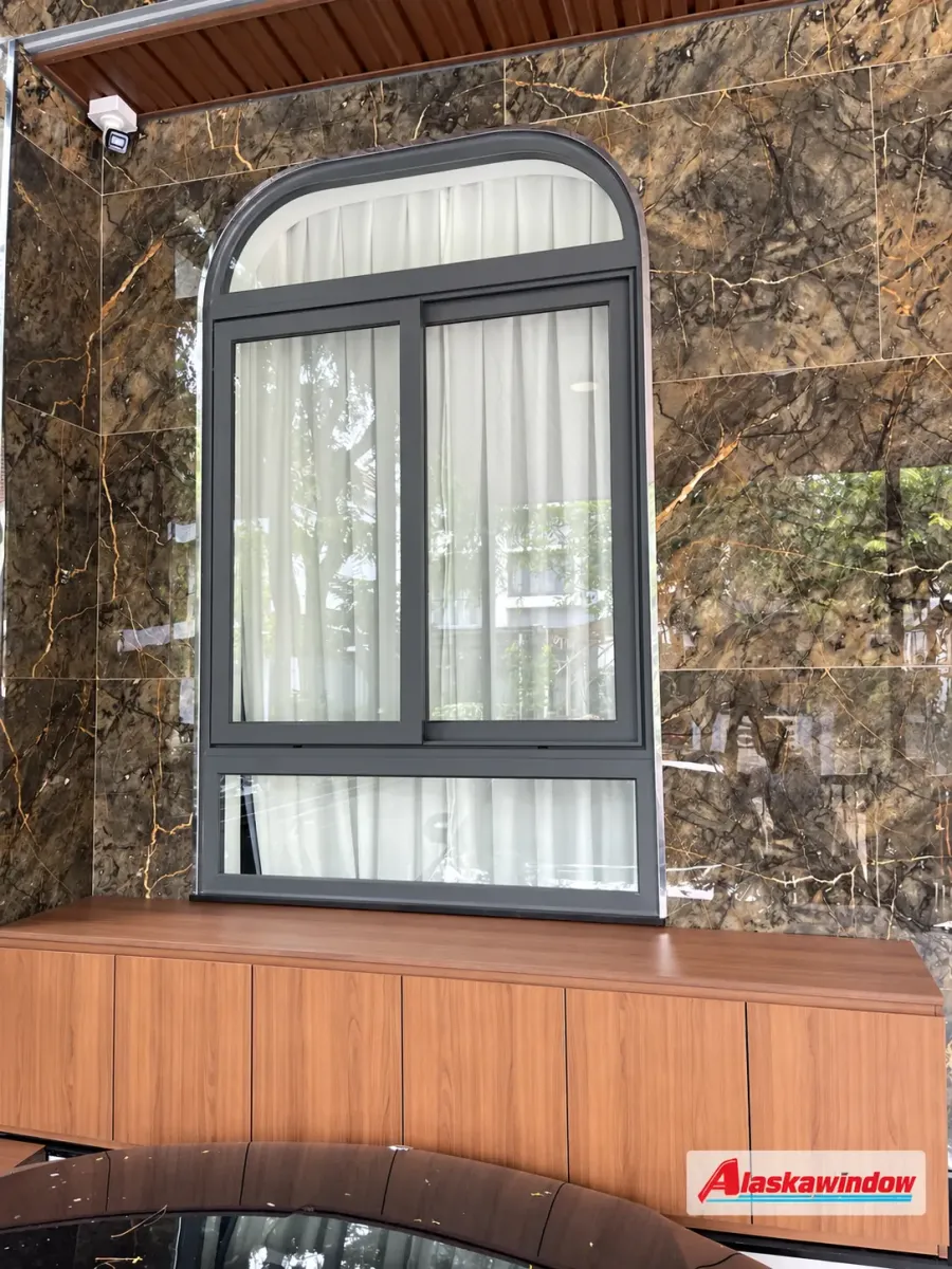 Cửa sổ nhôm xám ghi có ô vòm phía trên và rèm trắng trên nền tường ốp nâu