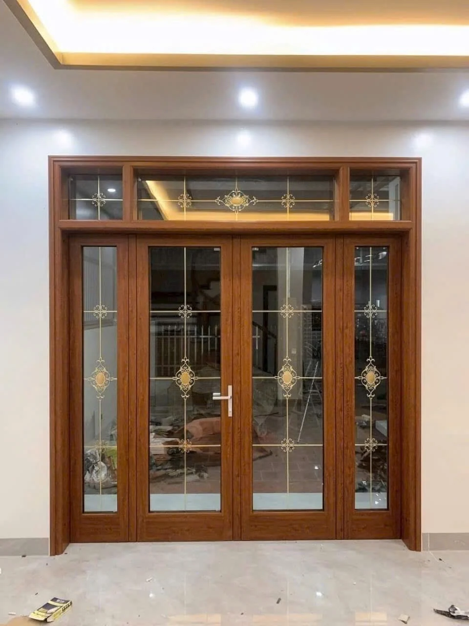 Bộ cửa màu vân gỗ nhiều ô kính hoa văn đã lắp, xung quanh còn vật dụng thi công